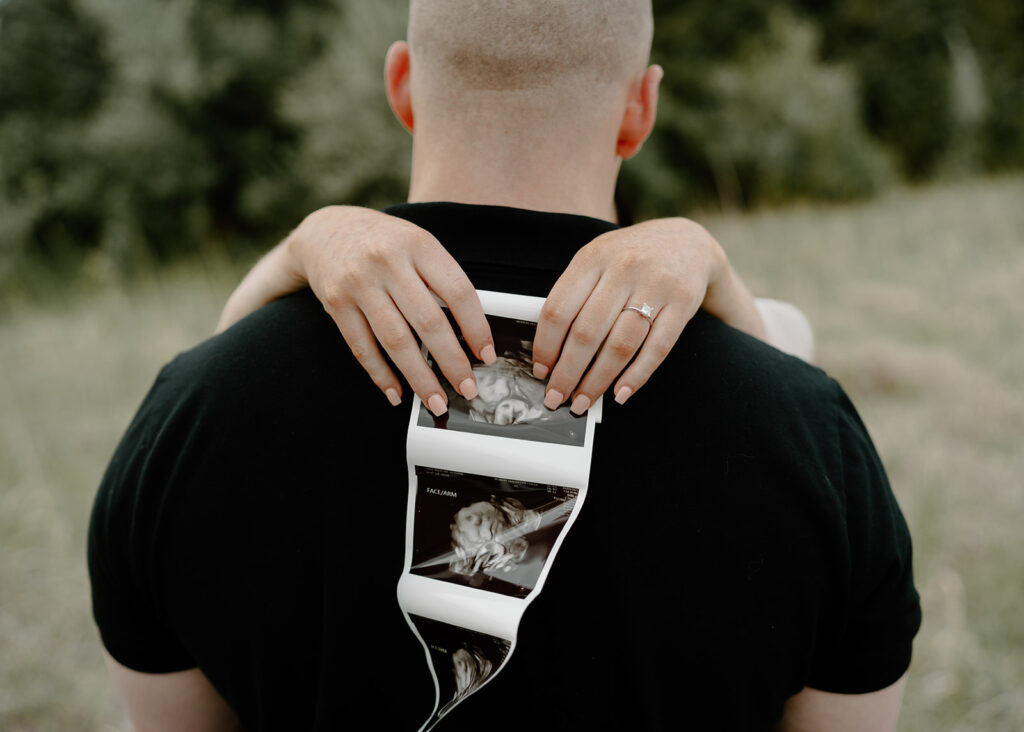 Woman holds ultrasound photo behind her partner's back for the family milestones collection session.